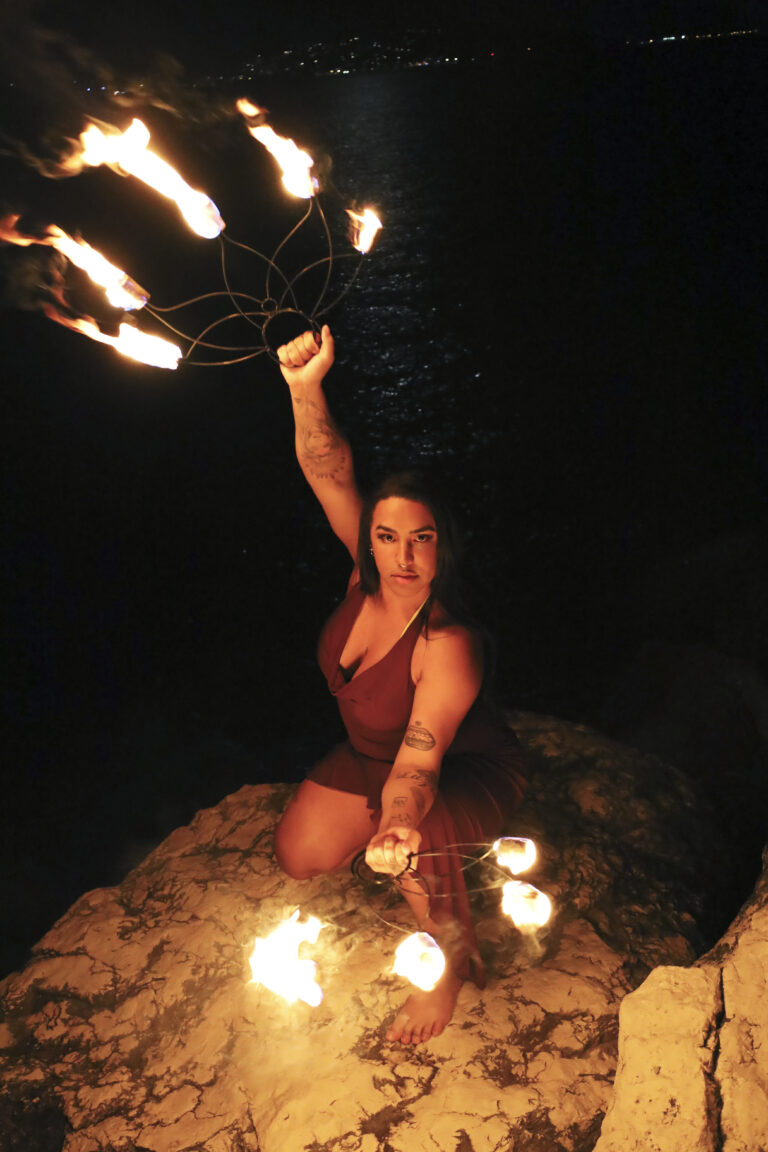 Photographie nocturne spectaculaire en plongée d'une performeuse (Bruna Taylor) agenouillée sur des rochers au bord de la mer. Elle porte une robe bordeaux et manipule deux éventails de feu enflammés, créant une chorégraphie lumineuse au-dessus d'elle et au niveau du sol. L'éclairage est assuré par les flammes et des lumières de ville lointaines sur l'horizon noir. Photographie de spectacle par BTarts.