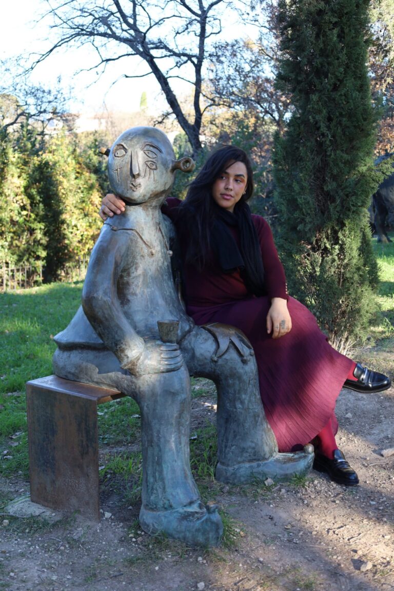 Plan moyen extérieur de Bruna Taylor en robe bordeaux plissée, collants assortis et mocassins noirs, posant assise sur le banc métallique d'une sculpture en bronze d'un personnage stylisé Picasso tenant un verre. Elle pose son bras autour de la sculpture. Le décor est un parc ensoleillé avec des cyprès et des arbres dénudés en arrière-plan. Photographie par BTarts