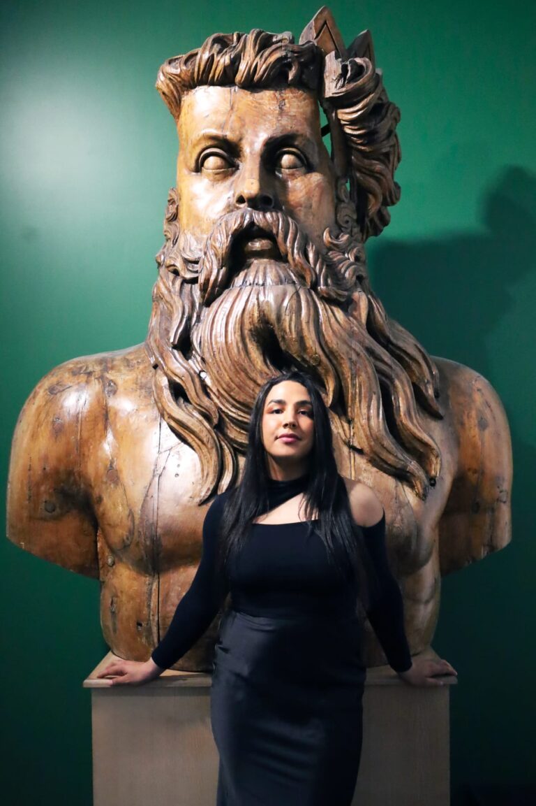 Portrait de face de Bruna Taylor, fondatrice de BTarts, portant un haut noir et une jupe en satin noir. Elle pose debout sur un socle en bois, encadrée par un imposant buste en bois sculpté d'un homme barbu aux cheveux longs, sur un fond de mur vert vif.