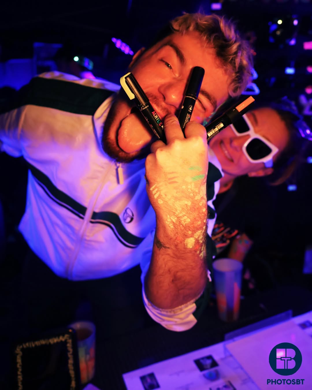 Photo dynamique de soirée sous lumière noire capturée par BTarts à Marseille . Un participant brandit des marqueurs Posca fluo avec la langue tirée, la main peinte. Un autre participant porte des lunettes de soleil fluo en arrière-plan. Ambiance ultraviolette intense.