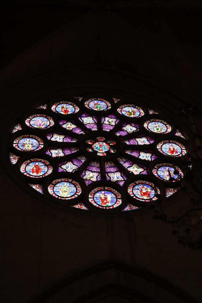 Vue en contre-plongée de la grande rosace en vitrail d'une cathédrale gothique. Le vitrail circulaire est complexe, composé d'un motif rayonnant de médaillons détaillés représentant des scènes bibliques et des figures religieuses en couleurs vives (bleu, rouge, or) sur un fond de pierre de voûte sombre et ombragé. Photographie d'architecture et patrimoine par BTarts.