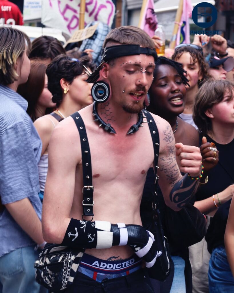 Portrait de street photography lors d'une manifestation techno, cadrage moyen d'un homme torse nu au look alternatif prononcé : nombreux piercings faciaux (nez, sourcils, joues), écarteurs d'oreilles massifs, tatouages sur le cou et les bras, collier à pointes et harnais à bretelles. Il porte des accessoires noirs avec des motifs d'ancre et un sous-vêtement 'ADDICTED' visible. Foule compacte et banderoles floues en arrière-plan. Photo par BTarts (logo visible).