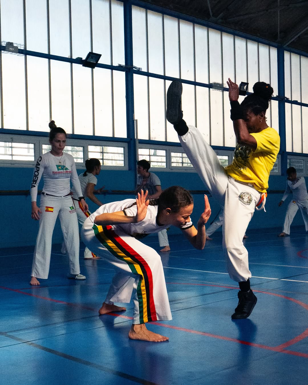 Photographie d'action lors d'un entraînement de Capoeira dans un gymnase au sol bleu. Au premier plan, une femme en abadá blanc et ceinture jaune-rouge-vert esquive en position basse (cocorinha), tandis qu'une autre femme en t-shirt jaune et pantalon blanc effectue un coup de pied haut esquivé. D'autres pratiquants et instructeurs sont visibles en arrière-plan flou. Reportage sportif par BTarts.