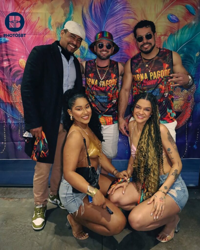 Photographie de groupe posée (plan moyen) de cinq amis lors d'un événement de musique brésilienne, devant un fond coloré imprimé de plumes et de masques. Deux femmes accroupies au premier plan et trois hommes debout derrière elles, tous souriants. Ils portent des t-shirts et débardeurs colorés à motifs (mention 'Carna Pagode Vamos Sambar'), lunettes de soleil et accessoires. Photo par BTarts.