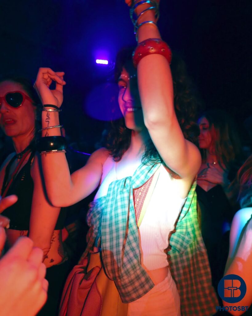 Photographie d'ambiance de clubbing nocturne, plan moyen d'une femme souriante, bras levés et poignets ornés de nombreux bracelets épais colorés (rouge, noir), dansant sous des lumières de scène magenta et violettes. Elle porte un top blanc et une chemise à carreaux turquoise et verte nouée autour de son buste. Photographie événementielle par BTarts