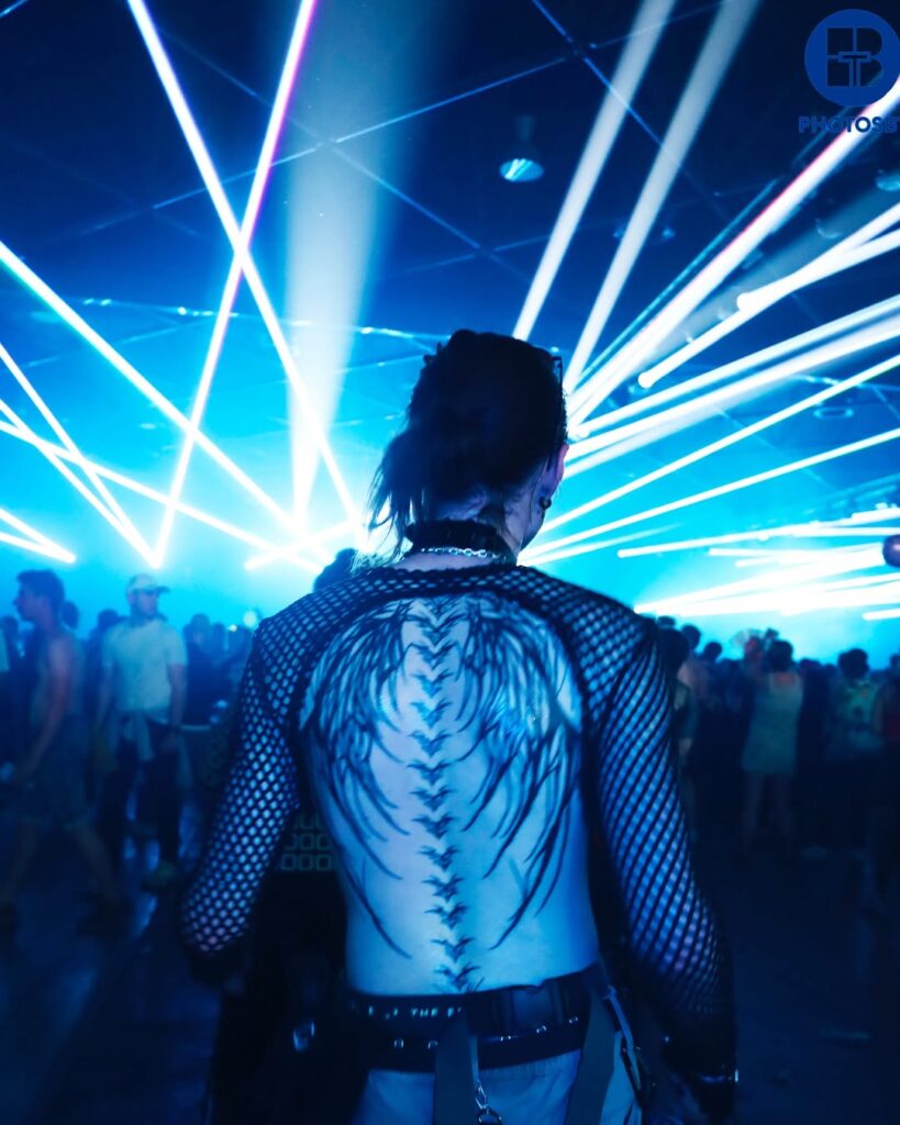 Photographie de plein pied artistique, plan de dos d'une personne à la silhouette fine et aux cheveux longs attachés, marchant dans un club ou un festival de nuit sous un ciel de faisceaux de lasers bleus et blancs intenses. La personne porte un haut en maille de filet noir qui révèle un tatouage d'ailes détaillé sur tout le dos. Foule et atmosphère nocturne floue en arrière-plan. Photographie par BTarts.