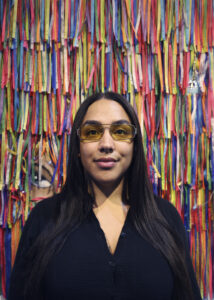 Portrait photographique de face de Bruna Taylor, fondatrice de BTarts, portant une chemise noire boutonnée et des lunettes de soleil aviateur à verres jaunes teintés. Elle pose devant un mur entièrement recouvert de centaines de rubans de vœux brésiliens (fitas do Senhor do Bonfim) multicolores et suspendus verticalement. Photographie artistique par BTarts.