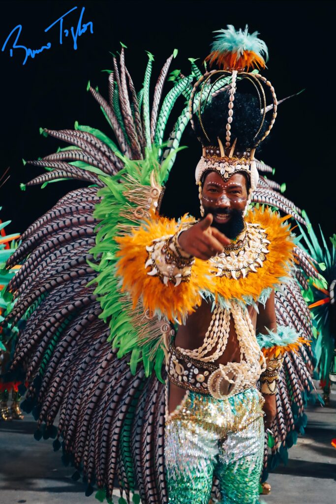 Portrait photographique d'un homme souriant, au premier plan, portant une coiffe de carnaval élaborée faite de plumes vertes et de faisan, ornée de coquillages cauris et d'une petite couronne à plumes turquoise. Il porte une collerette et une ceinture assorties de plumes orange et cauris, ainsi qu'un pantalon sequin brillant à motifs verts et or. Il pointe le doigt vers l'objectif sur un fond noir de défilé nocturne. Photographie événementielle par BTarts (signature 'Bruna Taylor' visible).