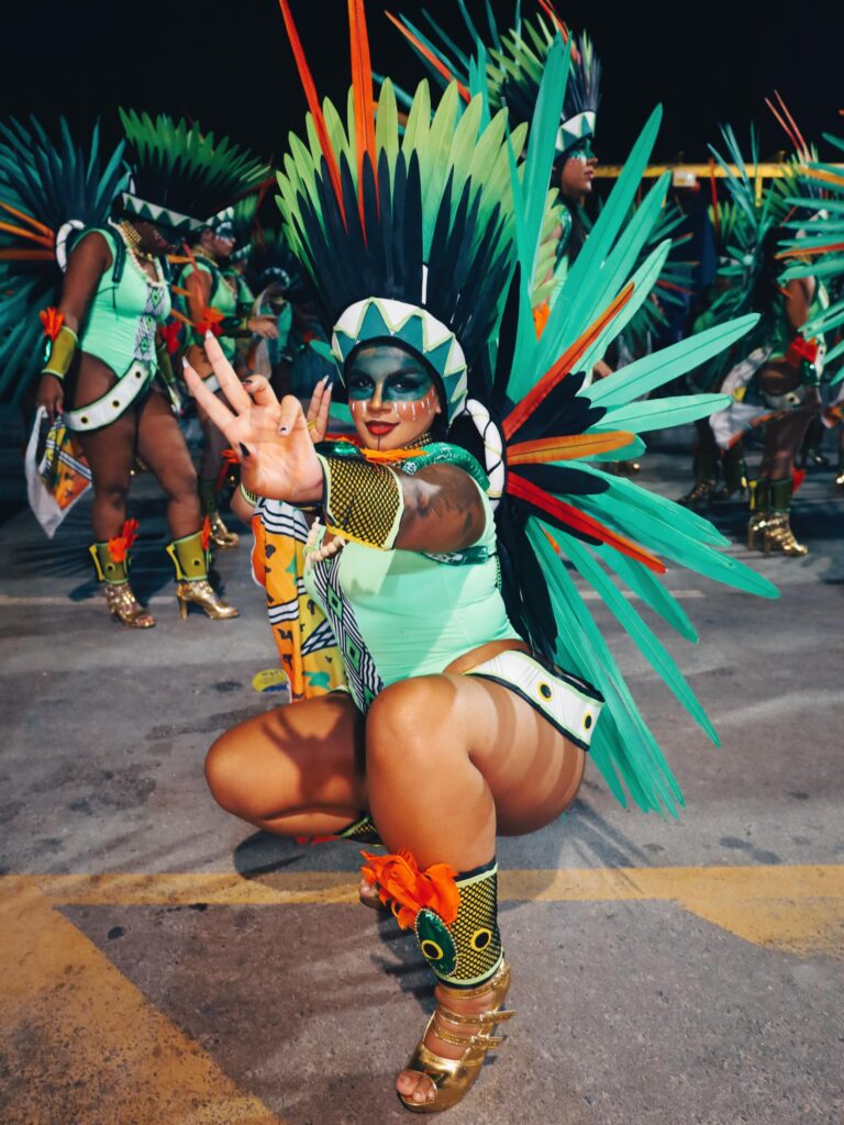 Photographie de plein pied d'une femme, accroupie sur le bitume, posant pour la caméra en costume de carnaval coordonné turquoise et or. Elle porte une grande coiffe de plumes turquoise et vertes, des bijoux de cheville coordonnés, et a le visage maquillé en turquoise. Elle lève une main, doigt en l'air. D'autres membres de la troupe en costumes similaires sont visibles en arrière-plan flou lors d'un défilé nocturne. Photographie de reportage par BTarts.
