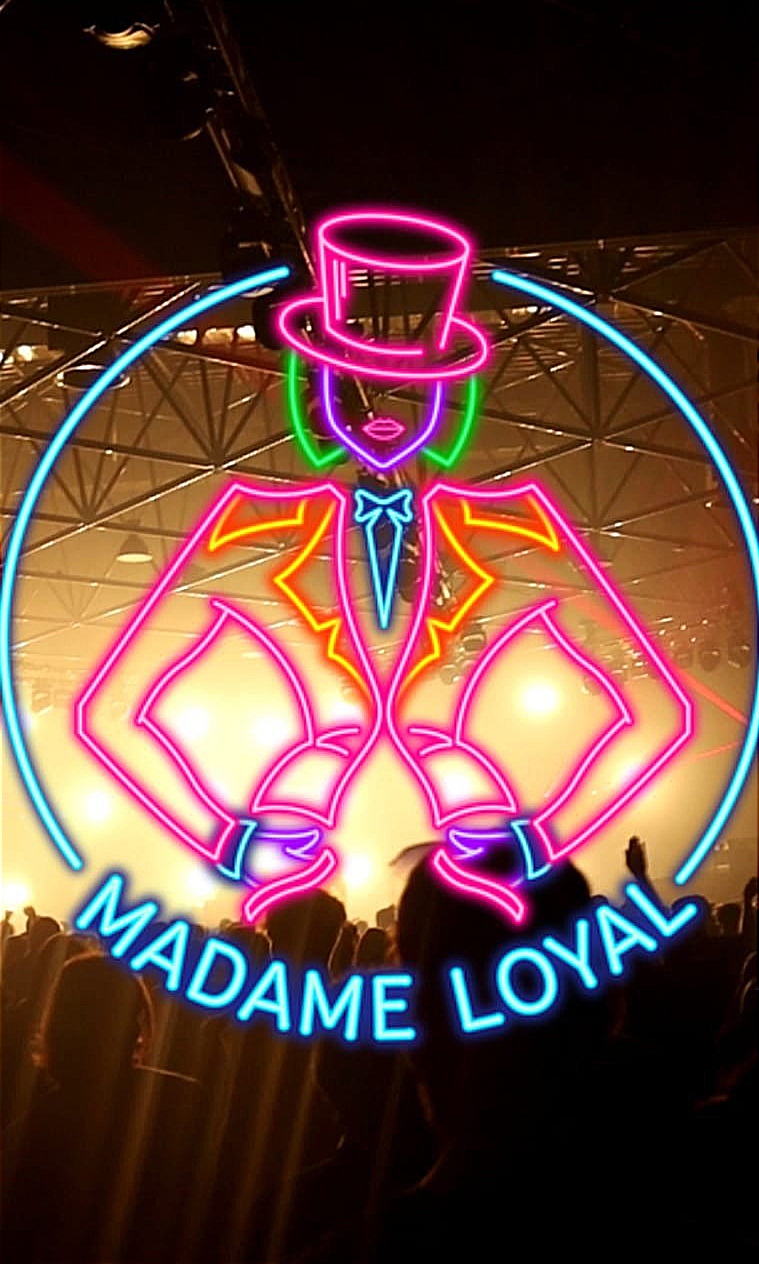 Capture d'ambiance d'un concert en salle avec foule, surmontée d'un logo en néon coloré (rose, bleu, vert) représentant une silhouette en chapeau haut-de-forme et costume, avec le texte 'Madame Loyal' en bleu. Photographie événementielle par BTarts