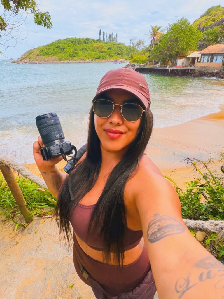 Portrait de Bruna Taylor, fondatrice de BTarts, avec un appareil photo professionnel type reflex et objectif à la main, sur une plage brésilienne avec collines et bungalows en arrière-plan.
