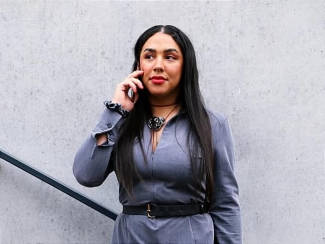 Portrait de Bruna Talor, avec de longs cheveux noirs, portant une combinaison grise, un collier floral noir, une ceinture noire à boucle et une montre, tenant un téléphone portable à son oreille droite et regardant vers la droite. Le fond est un mur de béton gris avec une rampe d'escalier noire sur la gauche.